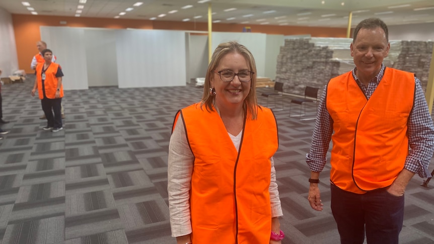 two people, wearing orange high-viz vests, standing in a big carpeted room 