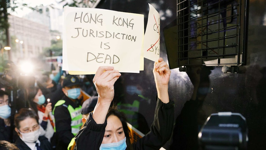 A woman wearing a mask holds a sign saying Hong Kong jurisdiction is dead with people gathered behind her.