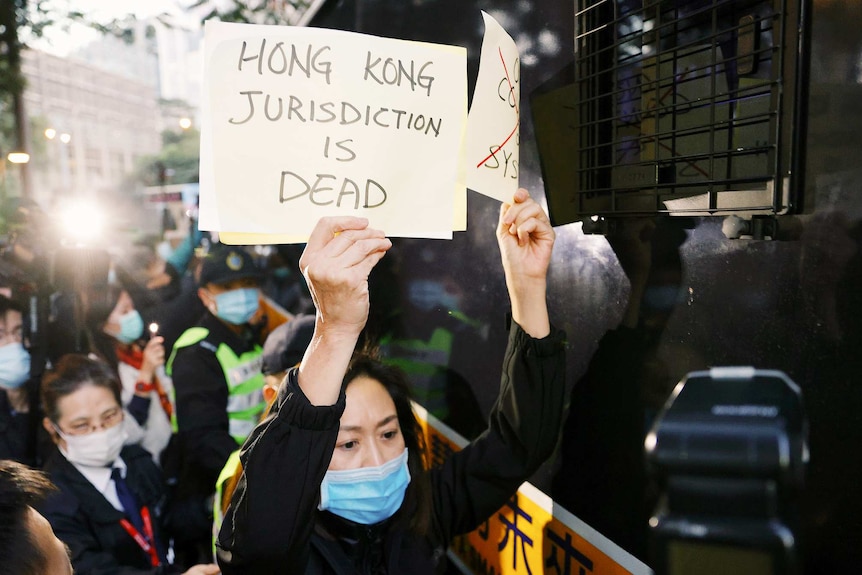 A woman wearing a mask holds a sign saying Hong Kong jurisdiction is dead with people gathered behind her.