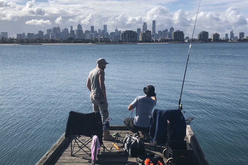 Two men sit on a dock fishing with the Melbourne skyline in the distance.