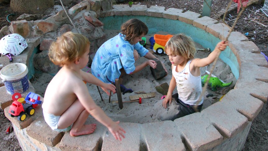 A toddler wearing only a nappy sitting on the edge of the sandpit watching his older sisters playing, holding a stick and a rock