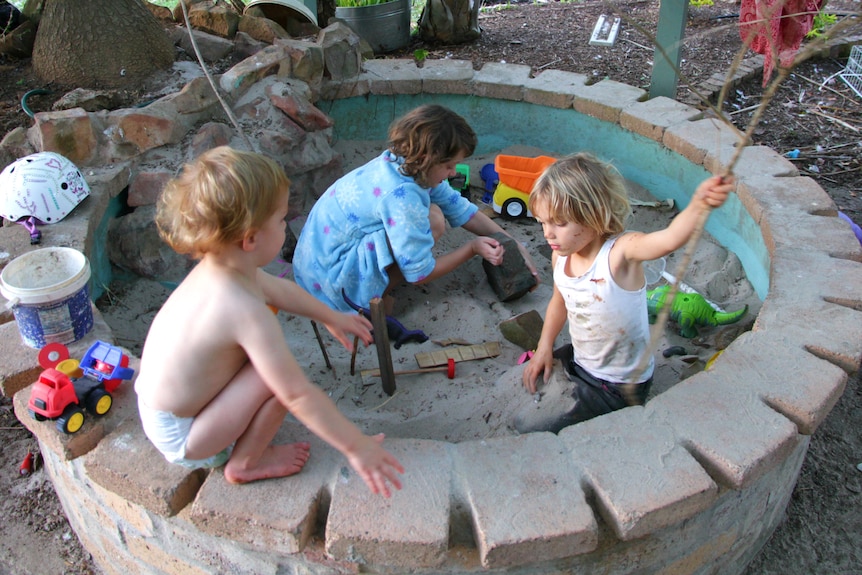 A toddler wearing only a nappy sitting on the edge of the sandpit watching his older sisters playing, holding a stick and a rock