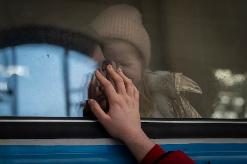 A hand reaches out to a train window, where a mother and crying child are sitting behind.