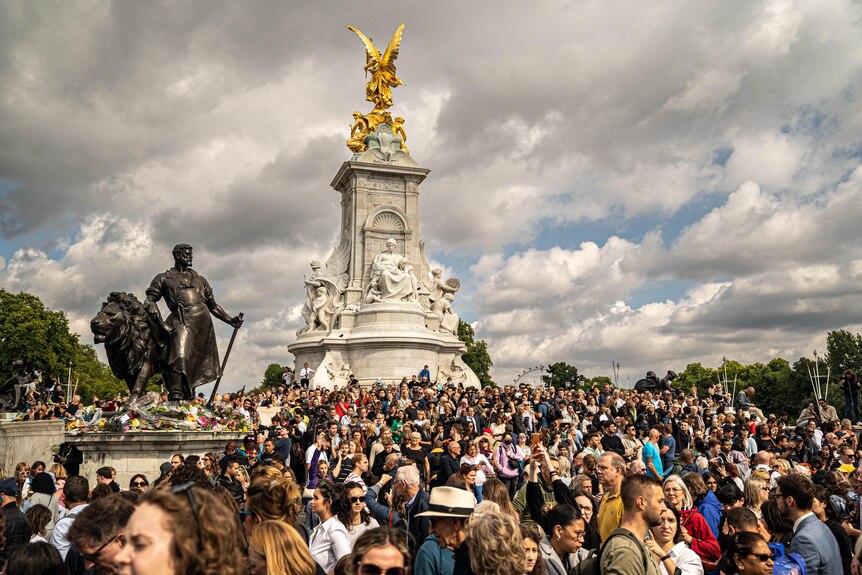 A group of people gathered around the marble and gold Victoria Tower under a cloudy sky