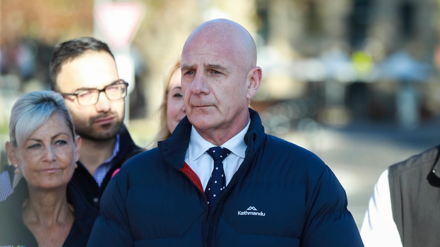 a bald man in a puffer jacket speaking to the media