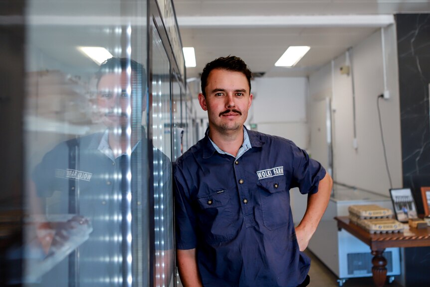 Man with moustache wearing blue shirt leaning on fridge inside butcher