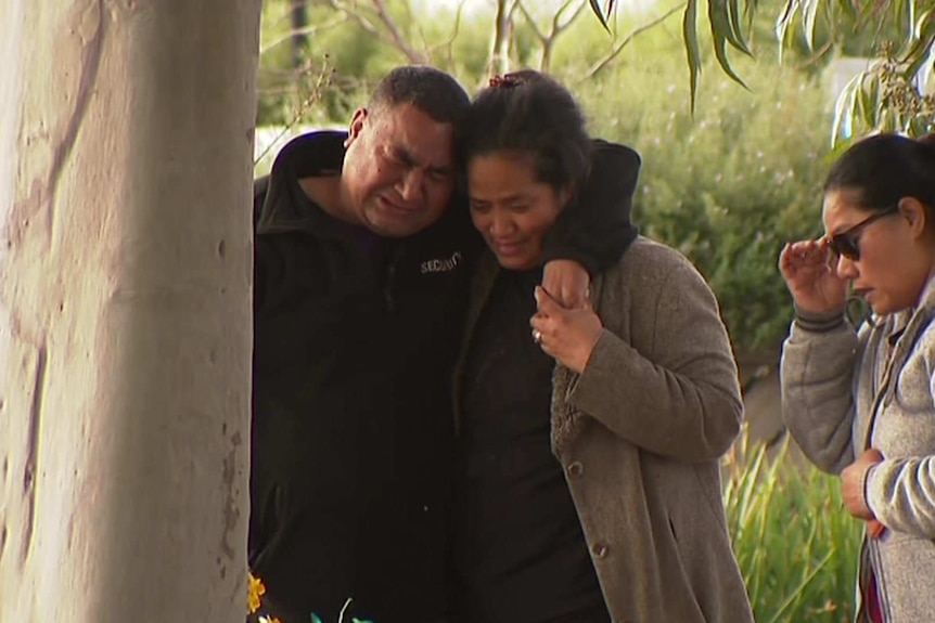 A weeping man and woman support each other next to a tree where they have laid flowers.