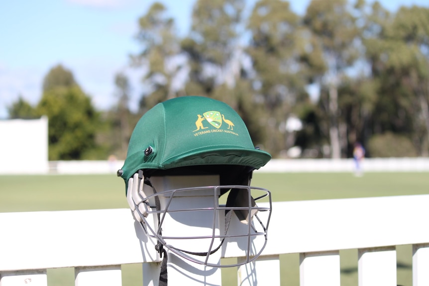 A green cricket helmet sits on a fence at a cricket oval.
