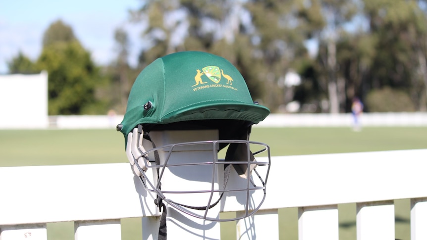 A green cricket helmet sits on a fence at a cricket oval.