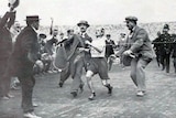 A man in shorts and a t-shirt crossing a finishing line while surrounded by men in suits and hats