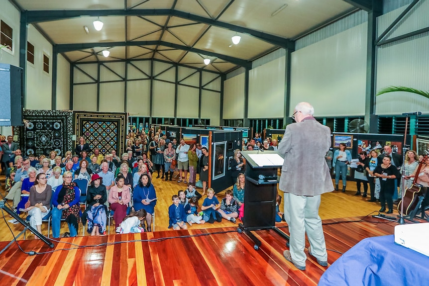 A man talks at an art exhibition opening in a large hall with people watching on.