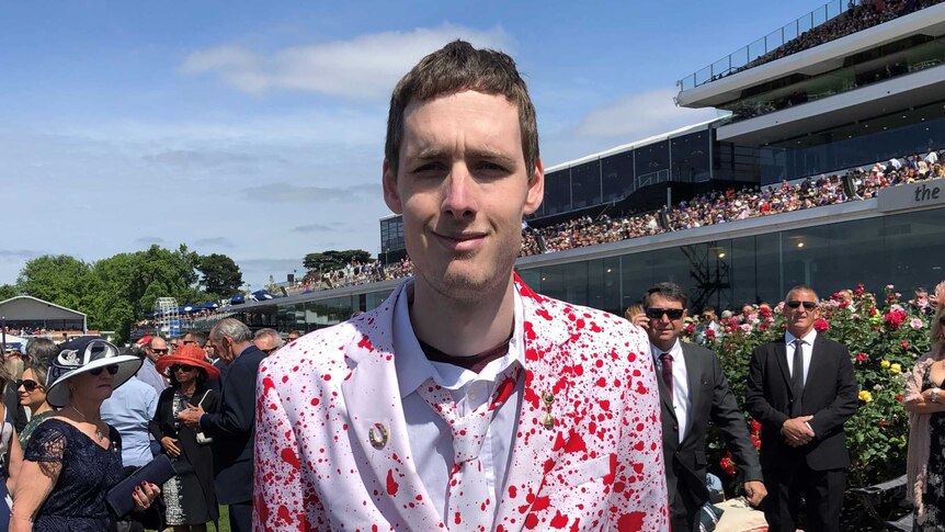 A man at the Melbourne Cup wearing a white suit with a blood splatter design.
