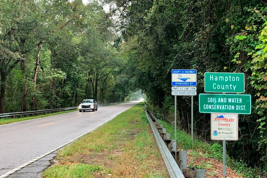 A sign welcomes travellers to Hampton County as a car drives down an empty road lined with trees on each side