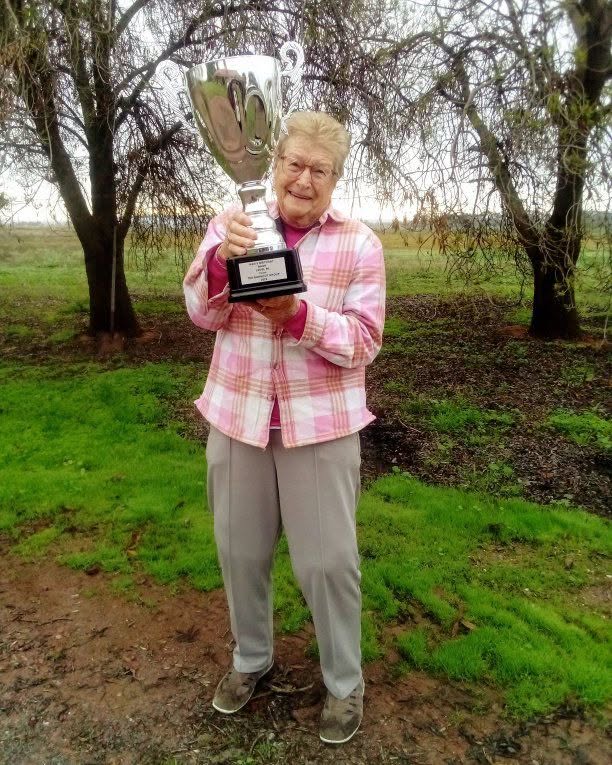 An older woman with glasses and blonde hair with a pink jacket stands smiling with a big silver trophy