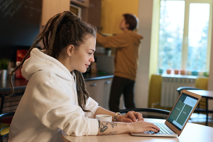 Woman with dreadlocks works with laptop on the kitchen bench while male puts something in cupboards