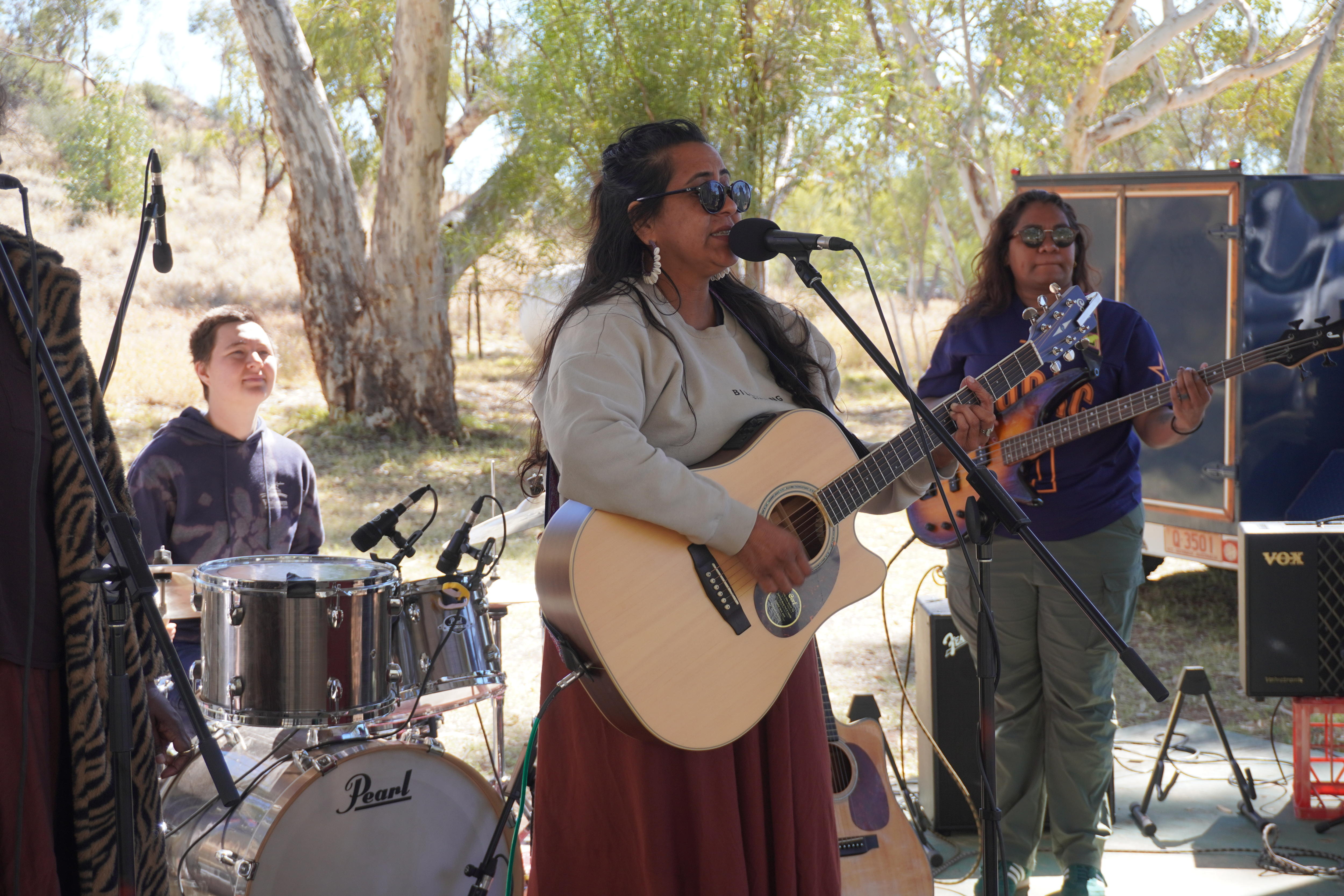 A woman sings into a microphone while playing a guitar. Behind two other women play drums and a guitar.