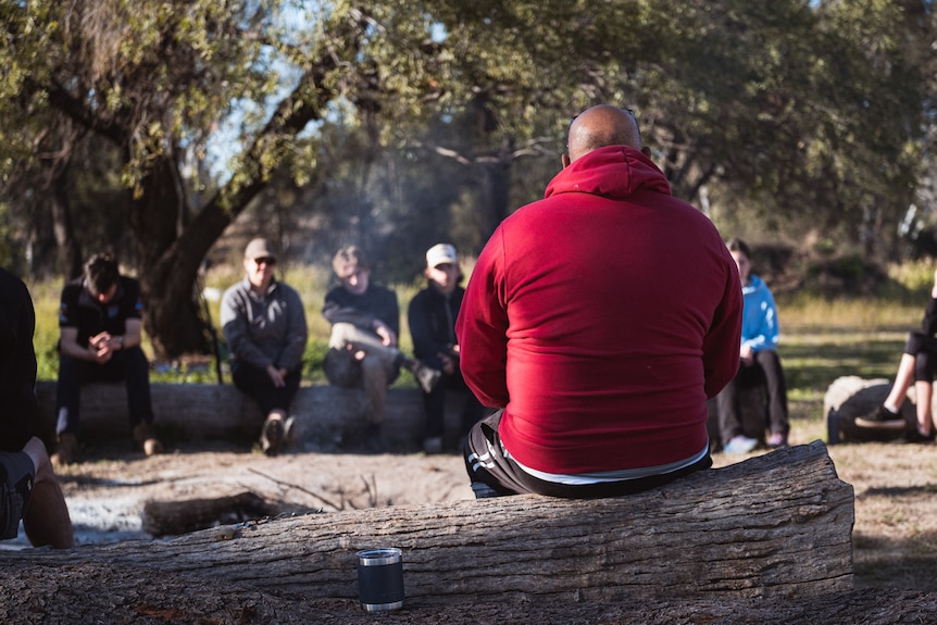 A man in a red jacket sitting on a log speaking to school children. 