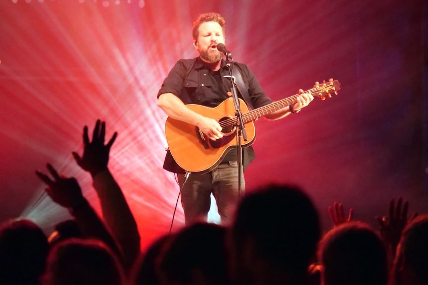 A man plays a guitar and sings into a microphone on a stage with reddish-pink lighting.