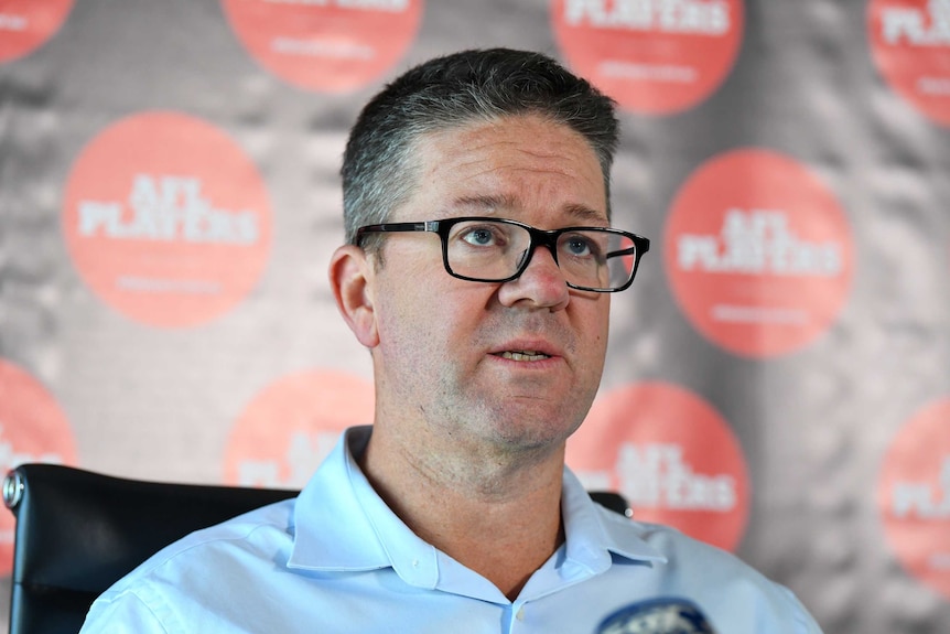 Man stands in front of microphone with a board behind him with AFL Players' Association logos on it.