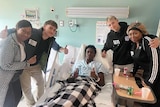 African American boy lies in hospital bed with four individuals crowded around him with thumbs up