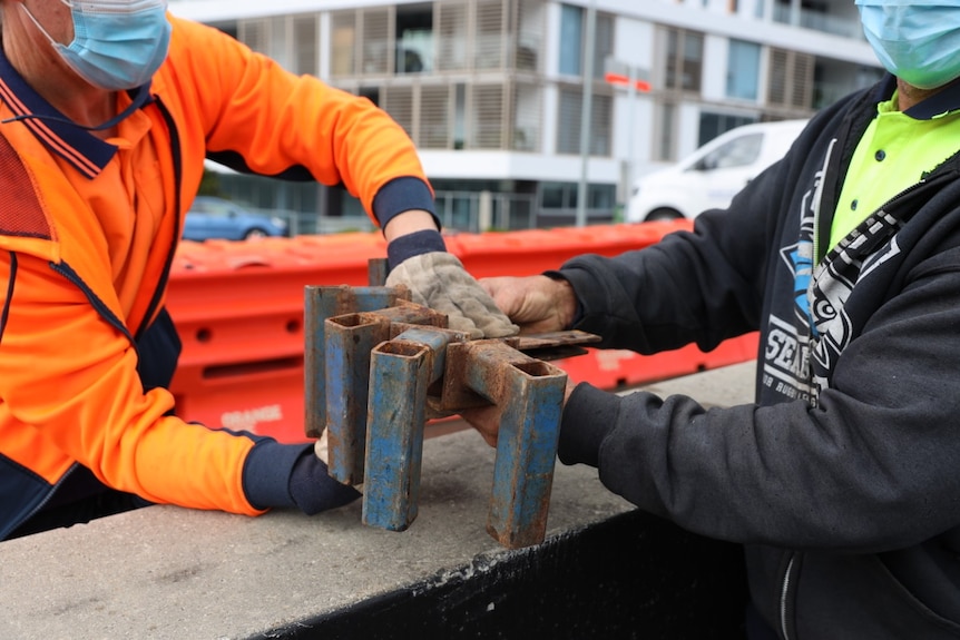 A man in orange high-vis receiving tools from a man in yellow high vis over border, orange barricade behind