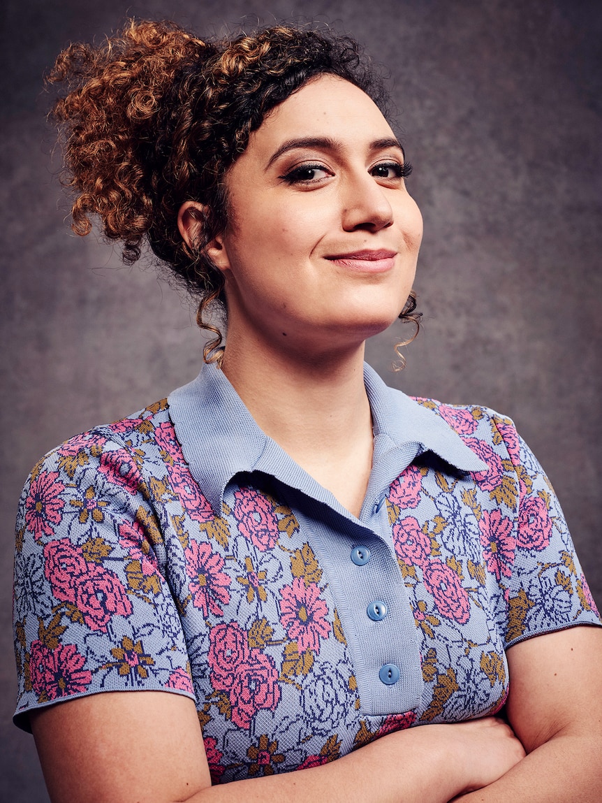 Rose Matafeo, her curly hair tied back, smiles proudly, with her arms folded 