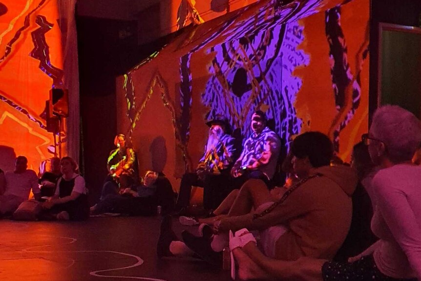 Gallery art space with red lighting, lighting paintings on walls, audience sitting on floor watching DJ at table on far left