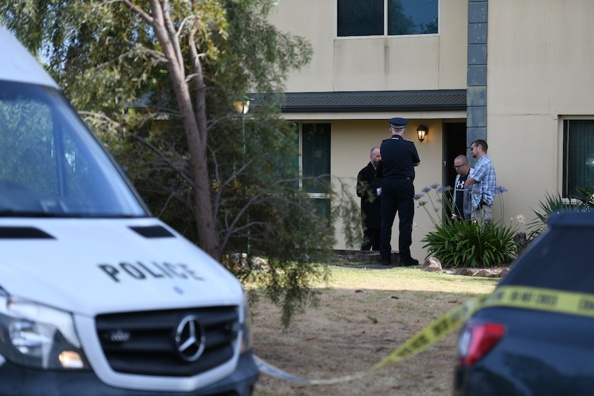 Police officers standing out the front of a two-storey house, behind police crime scene tape. 