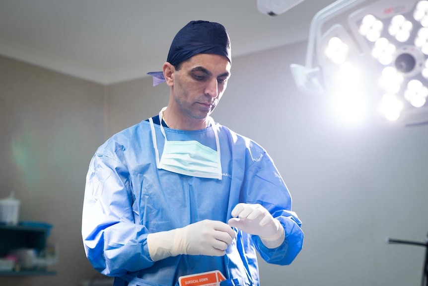 A surgeon in scrubs adjusts his gloves under bright lights