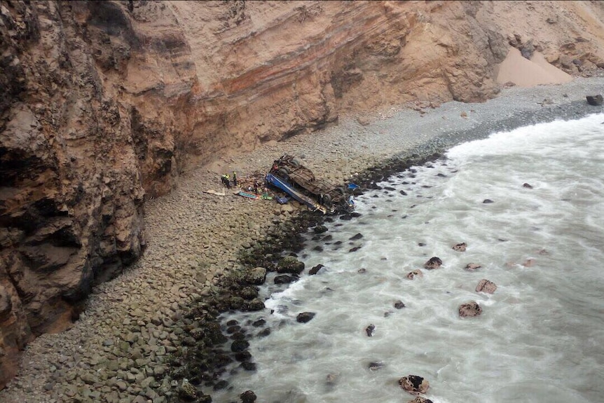 The wreck of a blue bus lies upside down on a beach near a tall, rocky cliff.