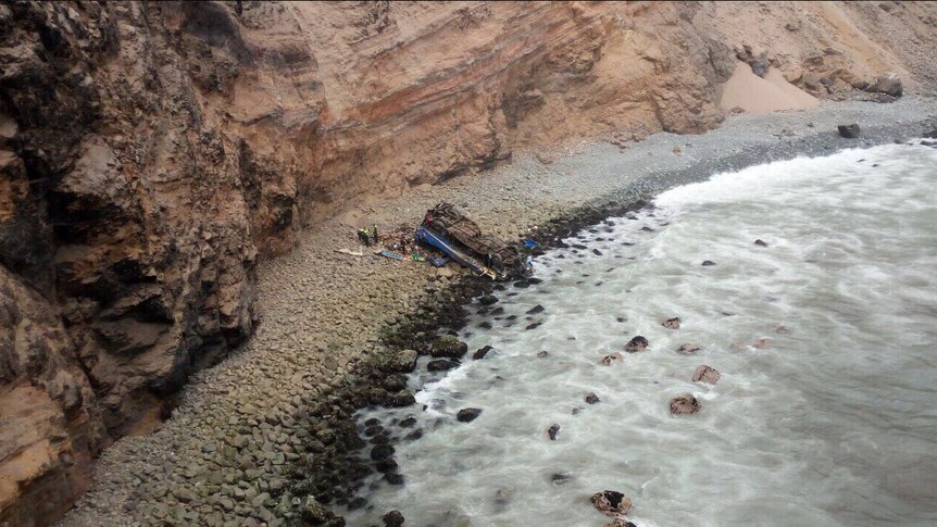 The wreck of a blue bus lies upside down on a beach near a tall, rocky cliff.