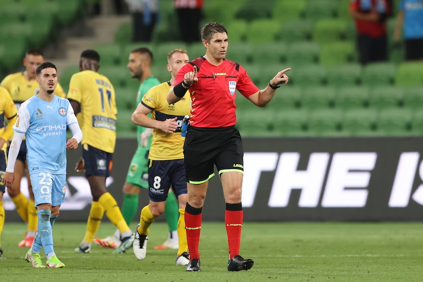 An A-League men's referee points with his fingers as he signals for a VAR check, as players walk after him looking on. 