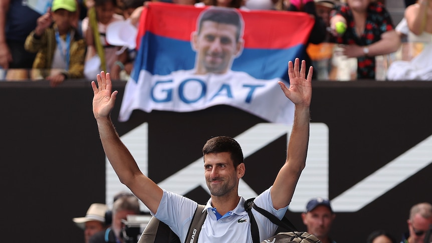 Novak Djokovic waves to Australian Open fans in front of a flag with his face and "GOAT" written on it as he leaves the court.