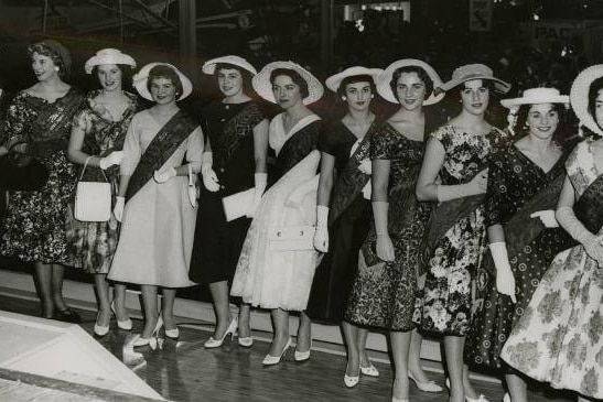 Black and white photo of showgirls in 1950s outfits, gloves and hats wearing sash ribbons.
