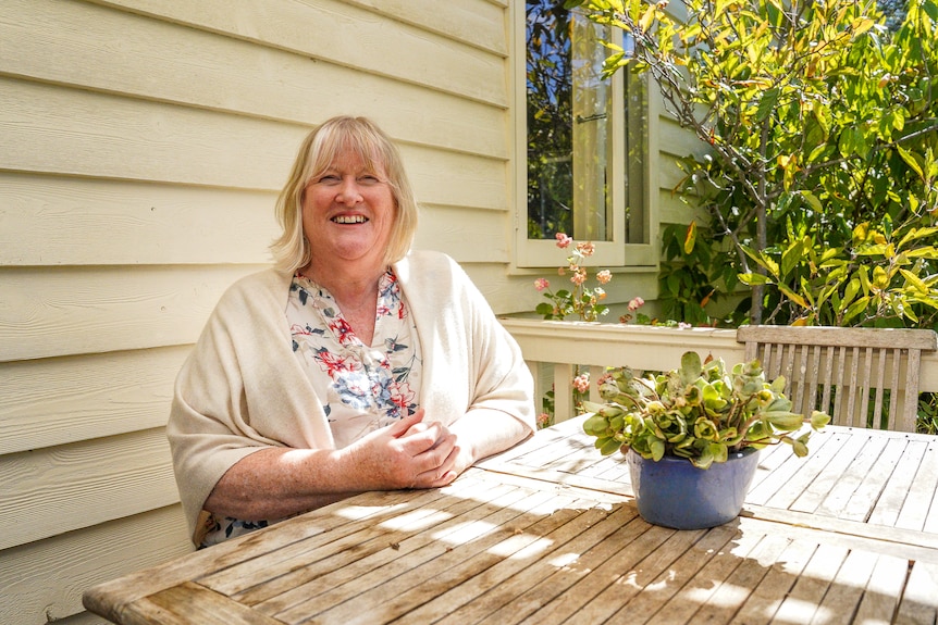 A woman sitting on a wooden deck of a weatherboard home wearing a floral shirt and white shawl.