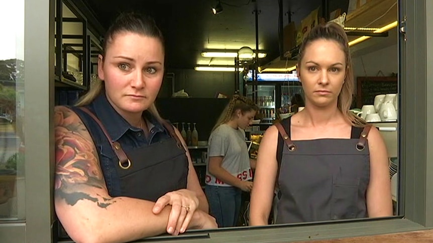 Two women wearing aprons stand behind a counter.