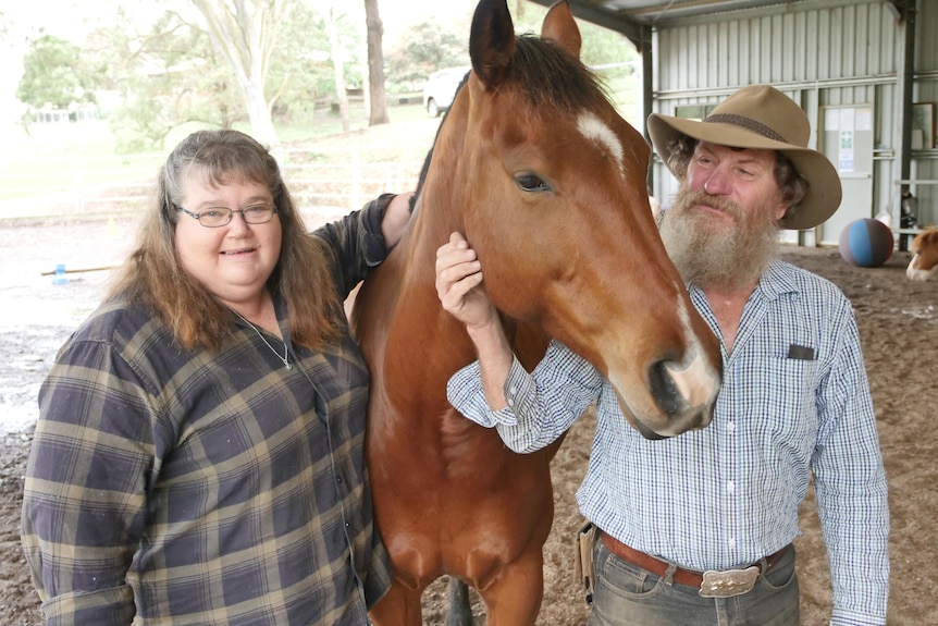 A couple stand with a Chessnut brown horse between them and their arms around him in a horse yard.