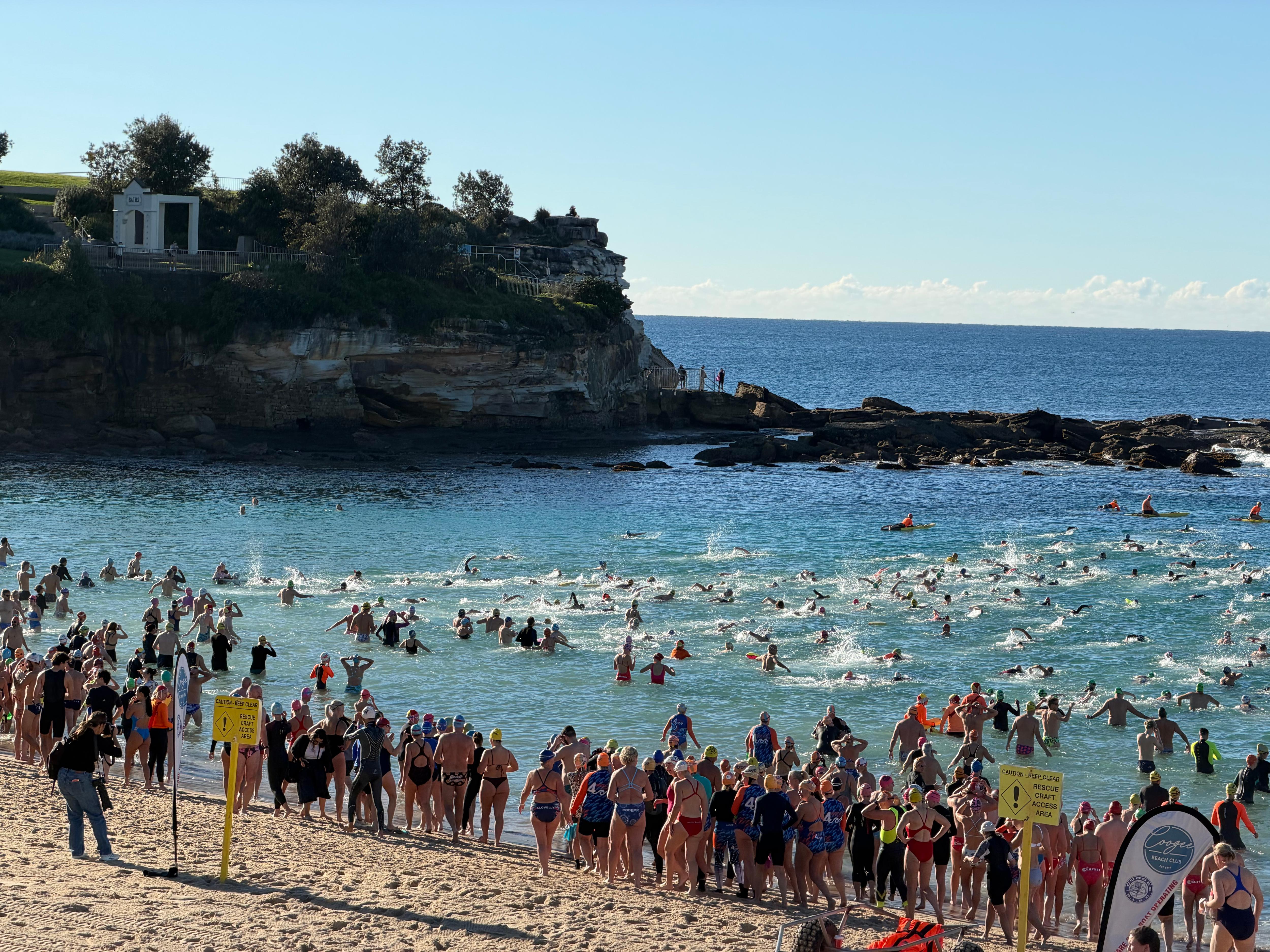 A large number of swimmers stand on the sand and swim out to sea with cliffs to their left.