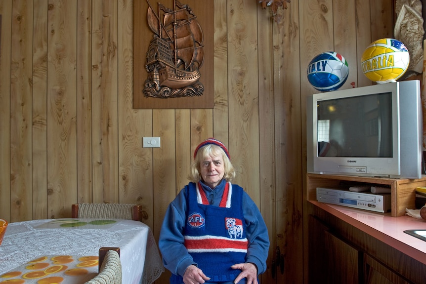 A woman wearing a knitted Western Bulldogs jumper and beanie sits in her wood panelled lounge room