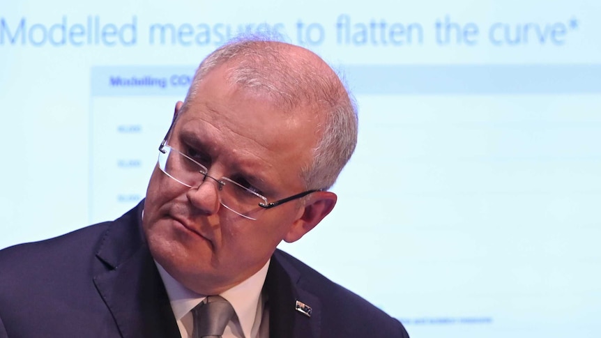 Scott Morrison tilts his head to the side during a press conference. A slideshow is visible on a screen behind him