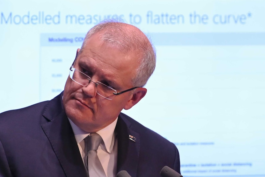Scott Morrison tilts his head to the side during a press conference. A slideshow is visible on a screen behind him