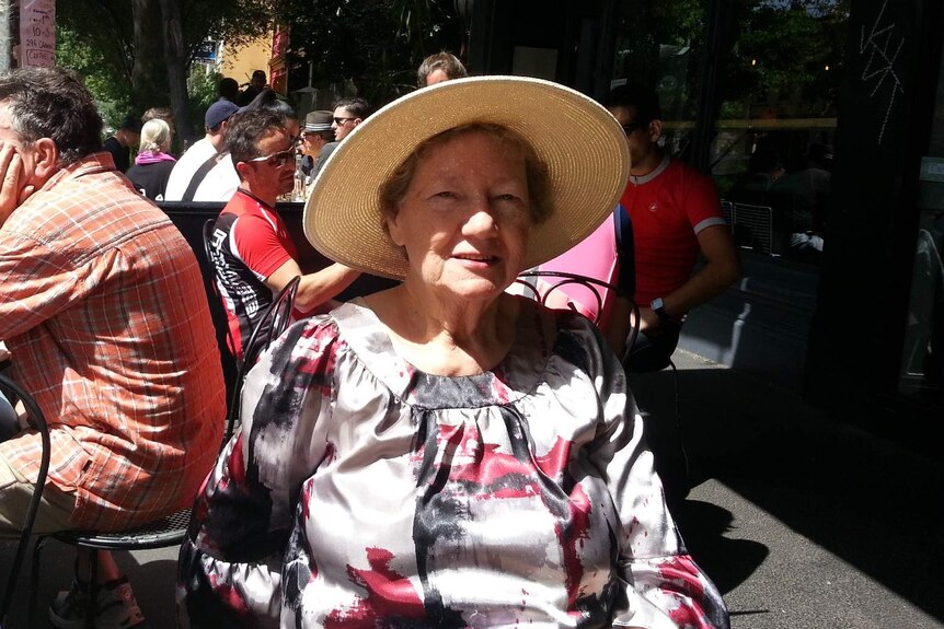 An elderly woman wearing a hat sits at an outdoor cafe in the sun, looking into camera.