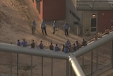 Inmates sit on a perimeter fence as police officers stand on the ground watching them. 