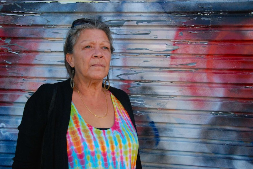 Older Indigenous woman standing against a colourful background.