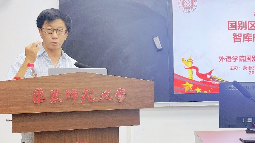 A man of Asian appearance wears glasses and stands speaking at a podium in a room, in front of a chalkboard and computer screens