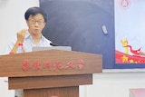 A man of Asian appearance wears glasses and stands speaking at a podium in a room, in front of a chalkboard and computer screens