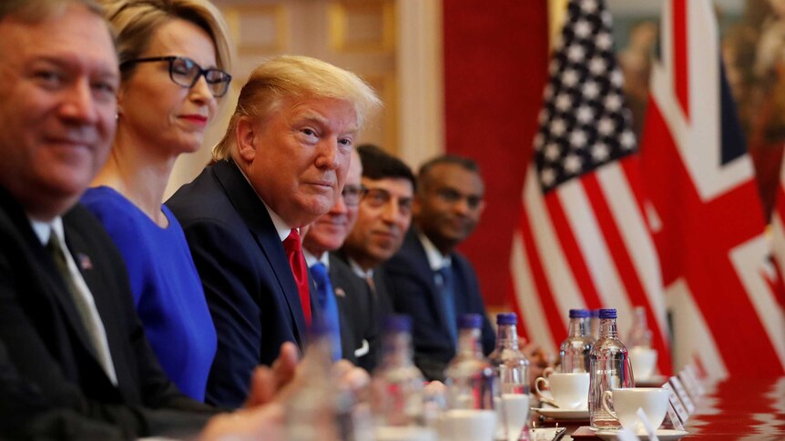 Donald Trump, seated next to other well-dressed people, looks down the length of a table. British and US flags sit on the table.