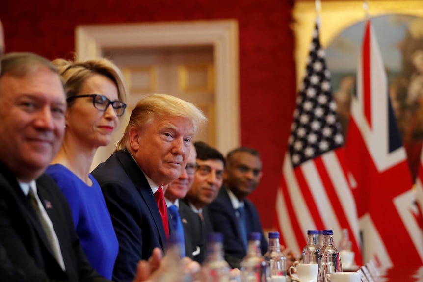 Donald Trump, seated next to other well-dressed people, looks down the length of a table. British and US flags sit on the table.