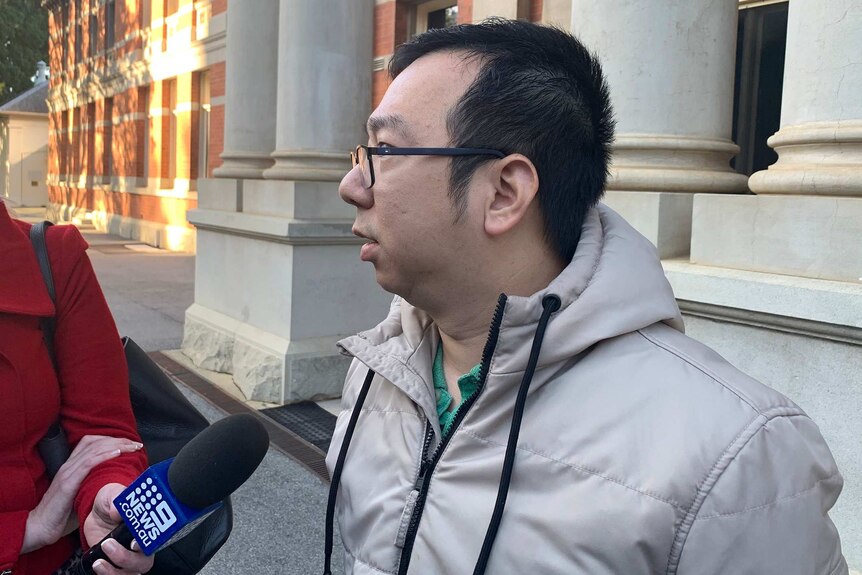 A side-on shot of a middle-aged man in glasses and a beige jacket talking into a reporter's microphone outside court.