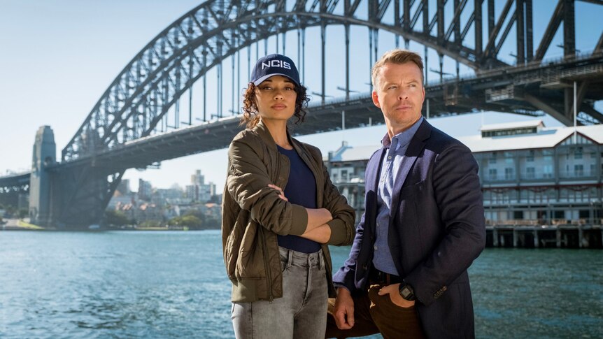 Two actors standing side by side in front of the Sydney Harbour Bridge with one wearing a cap with NCIS written on it